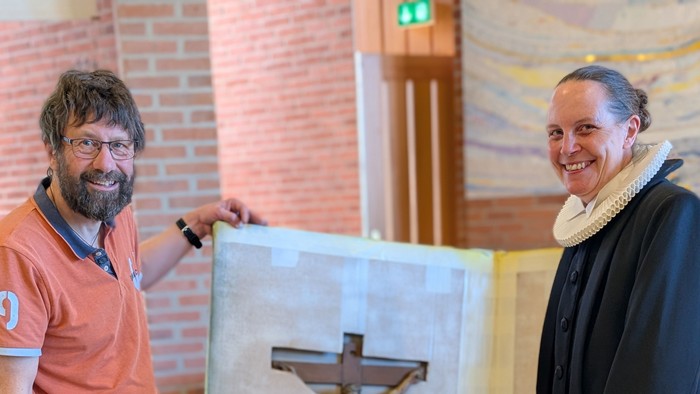 To personer poser med et krucifiks-kunstværk i en kirkelig omgivelse, den ene i afslappet påklædning og den anden i religiøst embedstøj.
