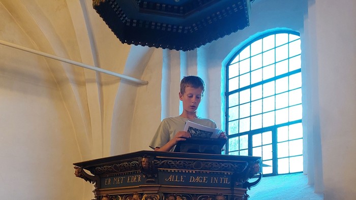 En ung dreng står ved et pulpet i en kirke og læser fra en bog