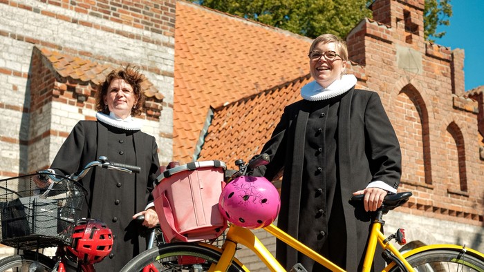 To kvinder i lange frakker poser med farverige cykler og kurve foran historiske murstensbygninger.