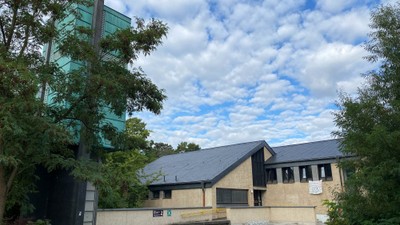 Moderne Gebäude mit Glasfassade und flachem Dach, umgeben von Bäumen und Himmel mit Wolken