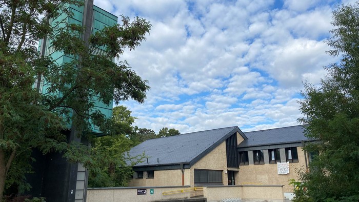 Moderne Gebäude mit Glasfassade und flachem Dach, umgeben von Bäumen und Himmel mit Wolken