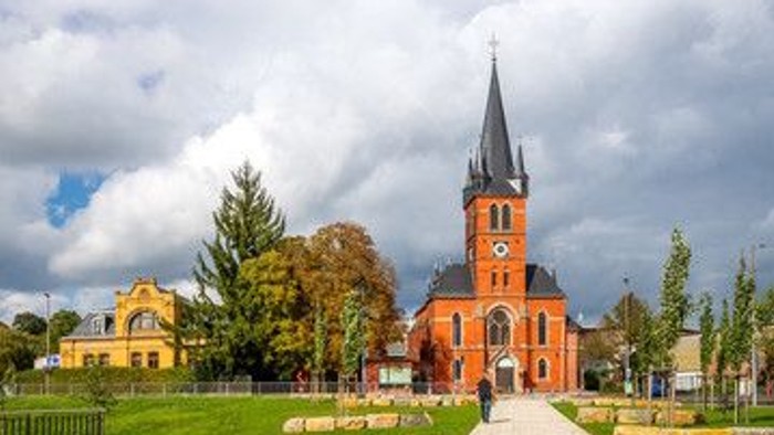 Eine malerische Kirche mit hohem Turm erhebt sich inmitten eines ruhigen, gestalteten Stadtplatzes unter dramatischem Himmel.