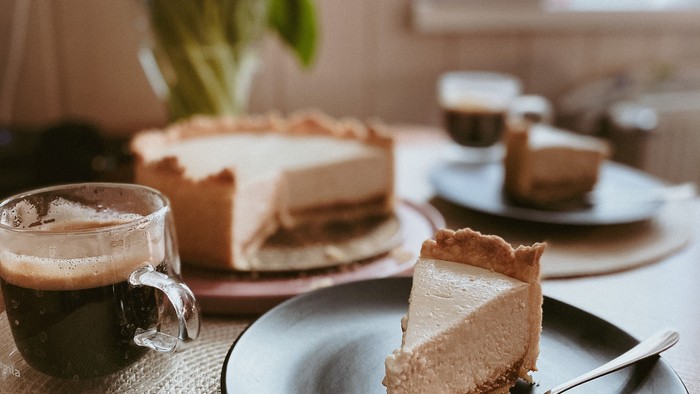 Ein Stück Kuchen auf einem Teller, daneben eine Tasse Kaffee und eine Vase mit Blumen