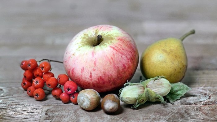 Auf einem Holztisch liegen ein rotes Apfel, eine gelbe Birne, rote Beeren und verschiedene Pilze