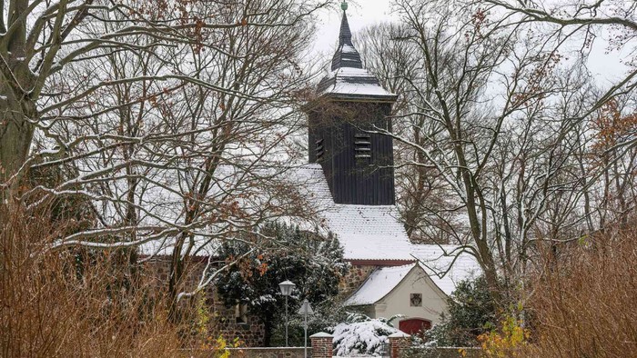 Schneebedeckte Kirche mit hohem Turm, umgeben von kahlen Bäumen.