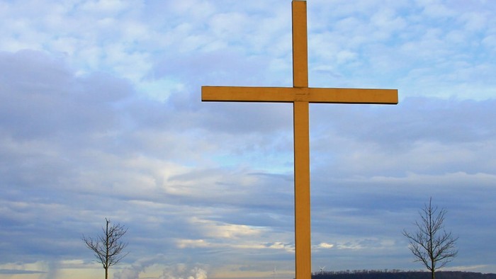 Ein großes Holzkreuz ragt hoch vor einem dramatischen, bewölkten Himmel mit kahlen Bäumen im Hintergrund auf.