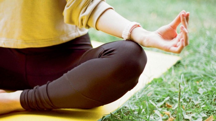 En person sidder i lotusstilling på en yoga-mat i græs.