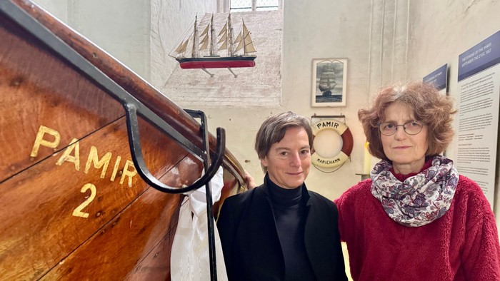 Zwei Frauen stehen neben einer Ausstellung mit einem hölzernen Boot, im Hintergrund sind ein Schiffsmodell und informative Tafeln zu sehen.