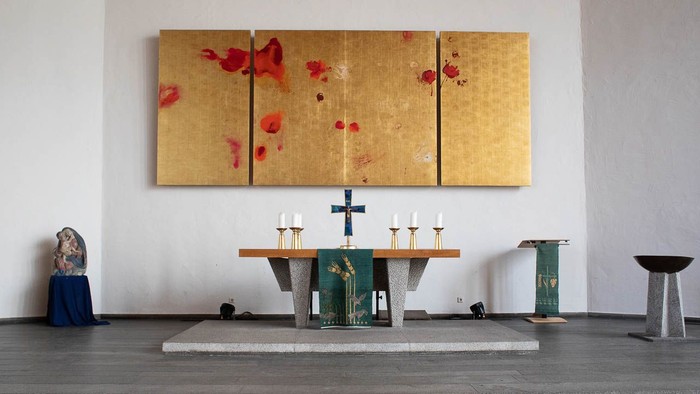 Ein Kircheninneres mit Altar, Kerzen und einem großen goldenen Bild mit roten Blumen an der Wand
