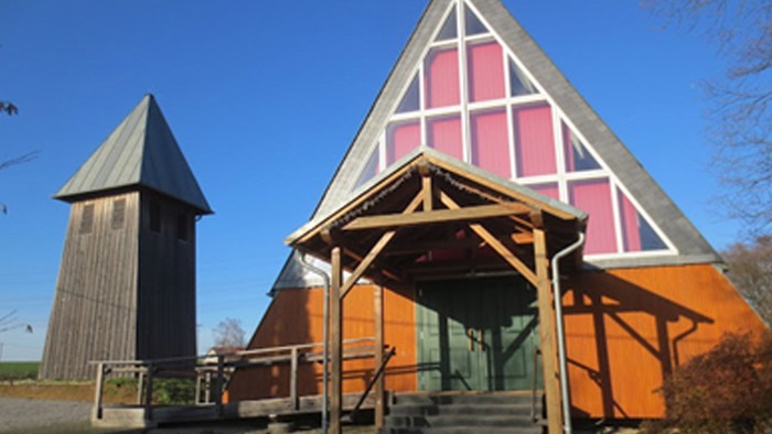 Ein modernes Holzhaus mit spitzem Dach und einer Treppe vor dem Eingang, daneben ein hohes, schlankes Gebäude.