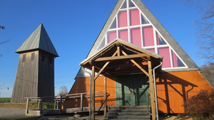 Ein modernes Holzhaus mit spitzem Dach und einer Treppe vor dem Eingang, daneben ein hohes, schlankes Gebäude.