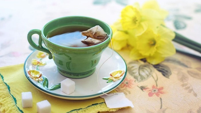 Eine grüne Teetasse mit Tee und Teebeutel auf einem Unterteller mit Zuckerstücken und einer gelben Blume daneben
