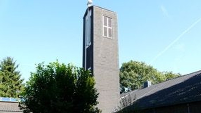 Ein hoher Backsteinkirchturm steht neben einem Gebäude mit grünen Bäumen und Bänken in einem sonnigen Innenhof.