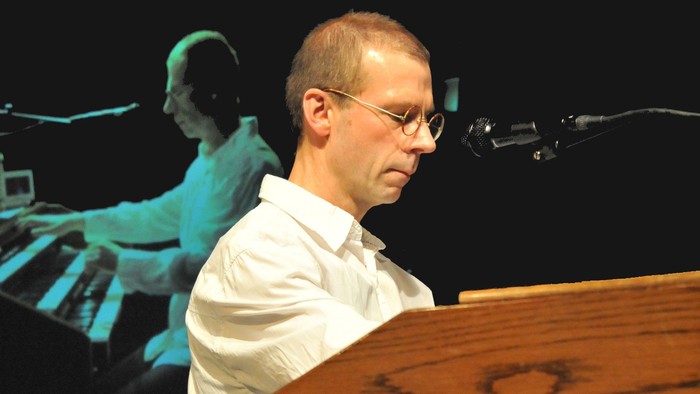 Mann steht hinter Pult und spricht in Mikrofon, im Hintergrund sitzt jemand am Keyboard