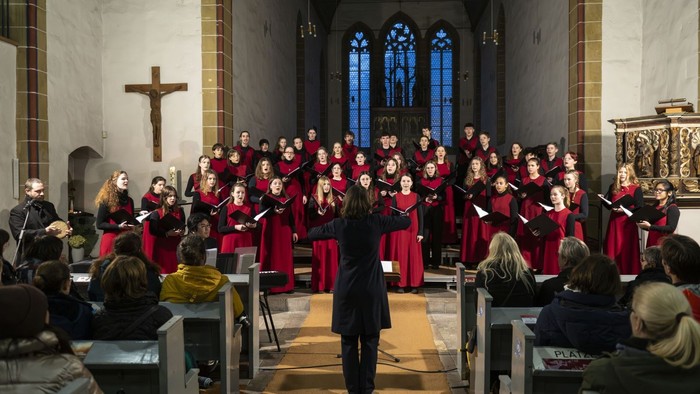 Ein Chor in roten Roben tritt in einer prächtigen Kirche auf, dirigiert von einem Dirigenten, während das Publikum in den Kirchenbänken sitzt.