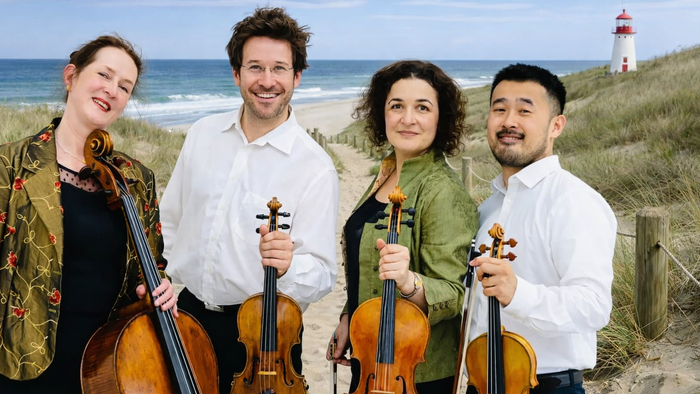 Ein Quartett von Musiker:innen mit Streichinstrumenten an einem Strand in der Nähe eines Leuchtturms.