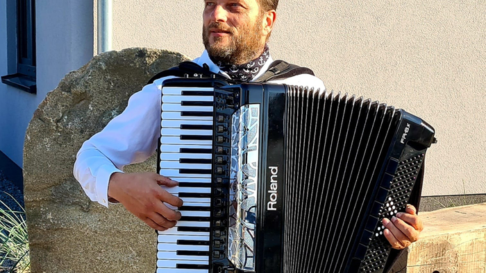 Mann spielt Akkordeon auf Holzbank vor Steinmauer