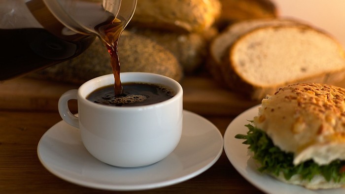 Kaffee wird in eine Tasse auf einem Holztisch gegossen, daneben ein Sandwich mit Salat und Käse sowie Brotlaib.