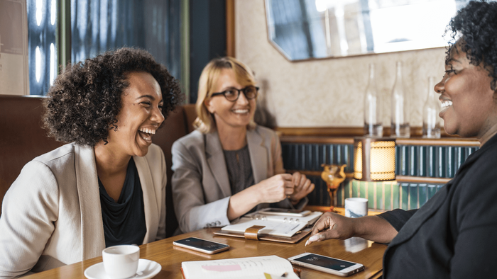 Drei Frauen lachen und unterhalten sich bei einer Kaffeepause im Büro.