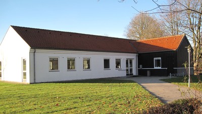 En hvid, langstraket hus med rødt tag og flere vinduer, placeret på en græsplæne med et mørkt, mindre hus ved siden.