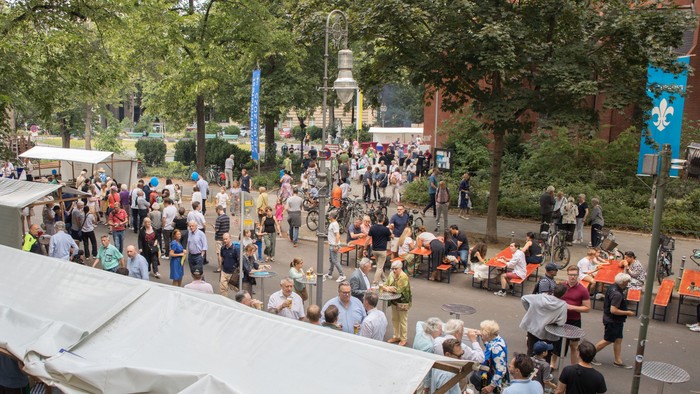 Belebter Freiluftmarkt mit zahlreichen Ständen und Menschen, die sich in einem baumbestandenen öffentlichen Bereich versammeln.