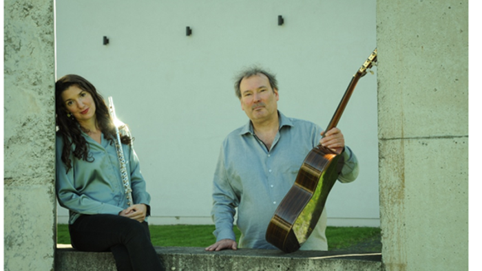 Ein Mann und eine Frau sitzen auf einer Betonmauer, er spielt Gitarre, sie hält eine Flöte.