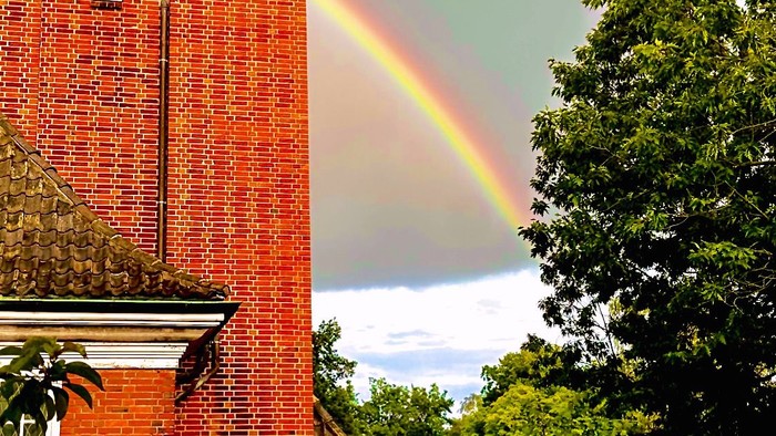 Links ein rotes Backsteingebäude, rechts ein Regenbogen über grünen Bäumen
