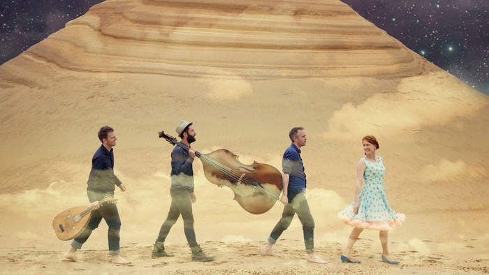 Fire mænd og en kvinde marcherer gennem ørkenen med instrumenter og en kasse.