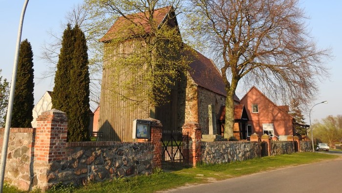Ein rotes Backsteingebäude mit spitzem Dach und hohem Tor, umgeben von Bäumen und einem Laternenpfahl.