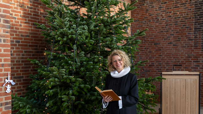 En person i religiøst tøj læser nær et pyntet juletræ i et udendørs område med murstensvægge.