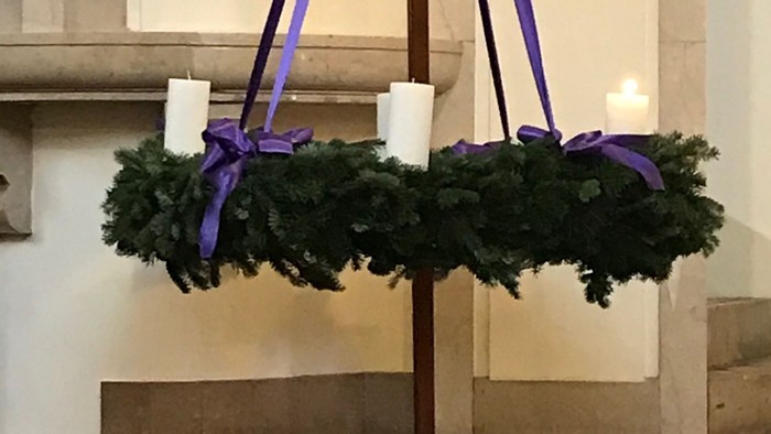 Ein Adventskranz mit drei brennenden Kerzen und lila Bändern vor einem steinernen Hintergrund