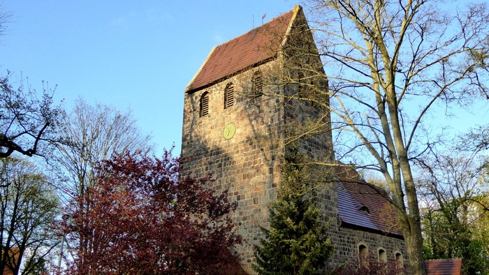 Alte Kirche mit hohem Turm und rotem Dach, umgeben von Bäumen und Büschen