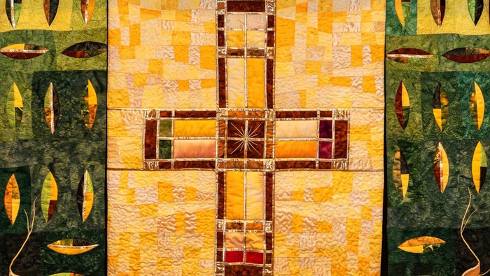 Das Bild zeigt ein farbenfrohes, religiöses Wandteppichwerk mit einem großen Kreuz in der Mitte.