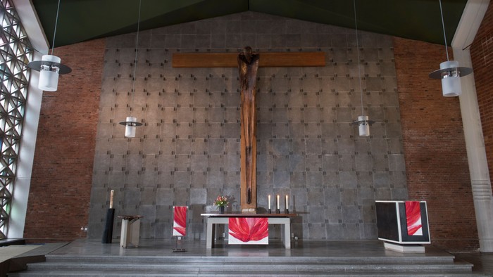 Die Kirche zeigt einen Altar mit Kreuz und Kerzen, flankiert von zwei roten Teppichen und einem Bildschirm.