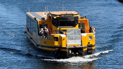En gult færge sejler gennem en byhavn med moderne og gamle bygninger.