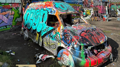 En bil dækket af graffiti i et forfalden område med murer og skralder.