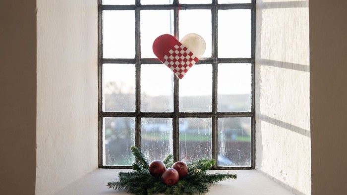 Hjerteformet pynt hænger i vinduet med grønne grene og røde bær nedenunder.