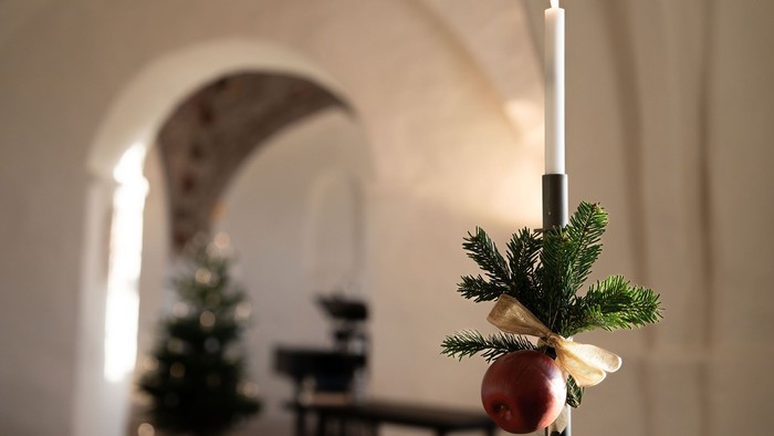 Stearinlys med grankviste og rød pynt i et rum med buer og juletræ.