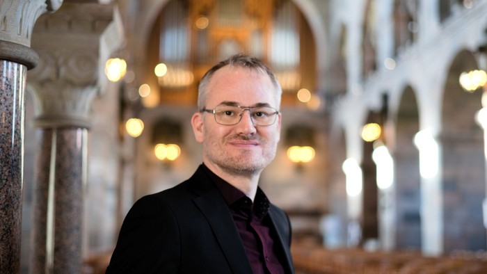 En mand i mørkt kostume og rød krage står i en stor, lys og pænt udsmykket kirke.