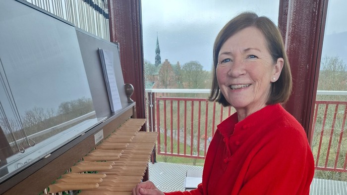 En person i en rød sweater spiller på et stort træinstrument, sandsynligvis et klokkespil, på en altan.