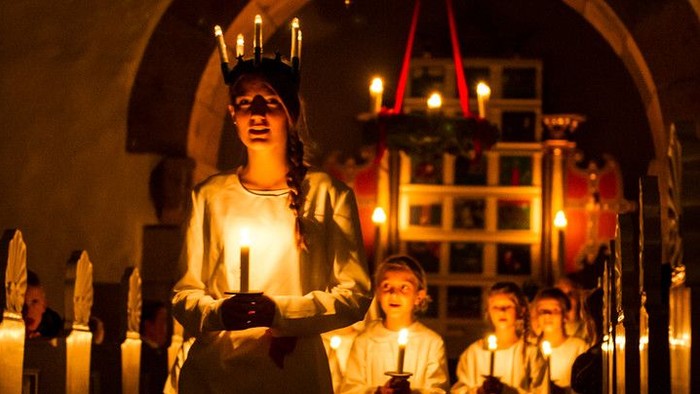 Børn iført hvide kåber, der holder lys, hvor den ene bærer en krone, står i et svagt oplyst område med buer og en juletræ.