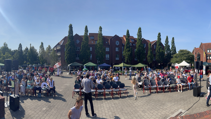 Menschen sitzen und stehen auf einem Kopfsteinpflasterplatz vor einem Gebäude mit Turm.