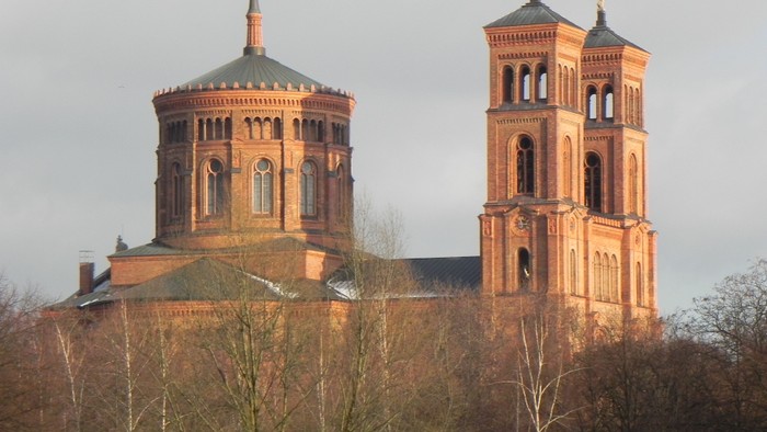 Zwei Türme einer Kirche mit spitzen Dächern und goldenen Akzenten, umgeben von Bäumen und einem bewölkten Himmel