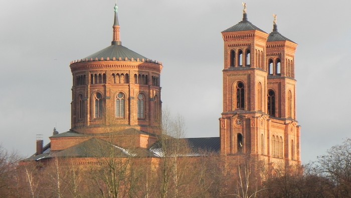Zwei Türme einer Kirche mit spitzen Dächern und goldenen Akzenten, umgeben von Bäumen und einem bewölkten Himmel