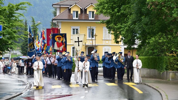 Messdiener mit Kreuz und Fahnen gehen vor einer Musikkapelle auf einer Straße entlang