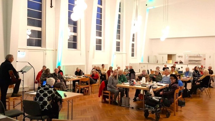 Eine Gruppe von Menschen versammelt sich in einem geräumigen Saal, sitzt an Tischen, während eine Person am Rednerpult spricht.