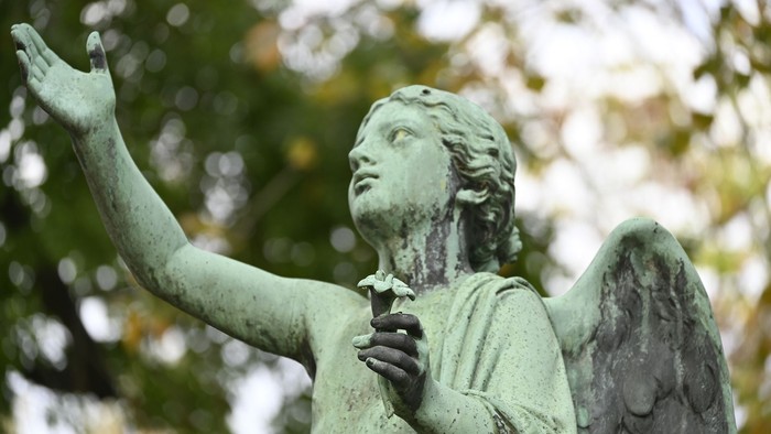 Engelsstatue mit erhobenen Armen und gefalteten Händen, umgeben von Bäumen