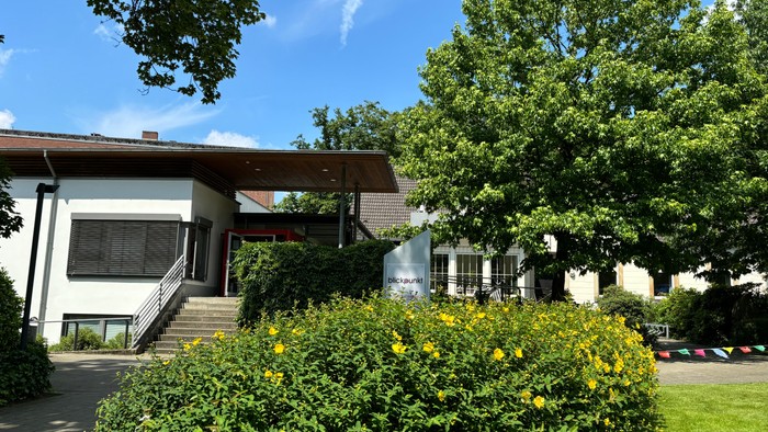 Hier sieht man das Gemeindehaus blick.punkt, ein weißes, flaches Gebäude mit großer Treppe und einer roten Tür.