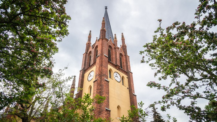 Kirchturm mit Uhr und Spitzhelmen, umgeben von Bäumen.