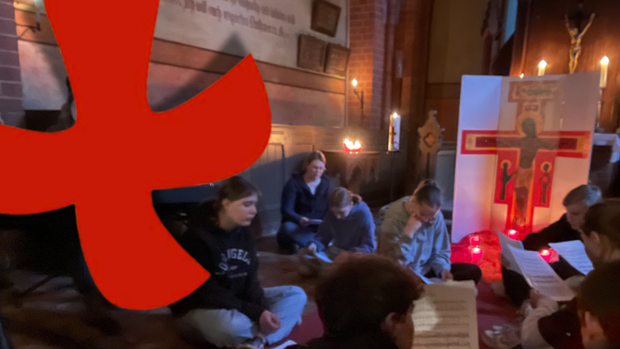 Personen sitzen in einer Kirche, ein großes rotes Kreuz ist prominent im Bild.